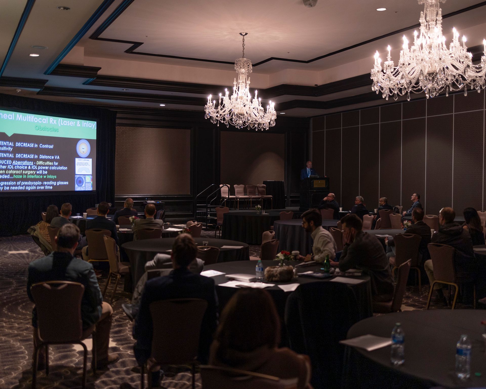 A group of people are sitting at tables in a conference room watching a presentation