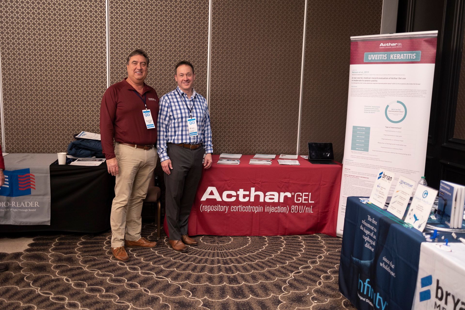 Two men are standing in front of a table with a red table cloth that says acthargel.