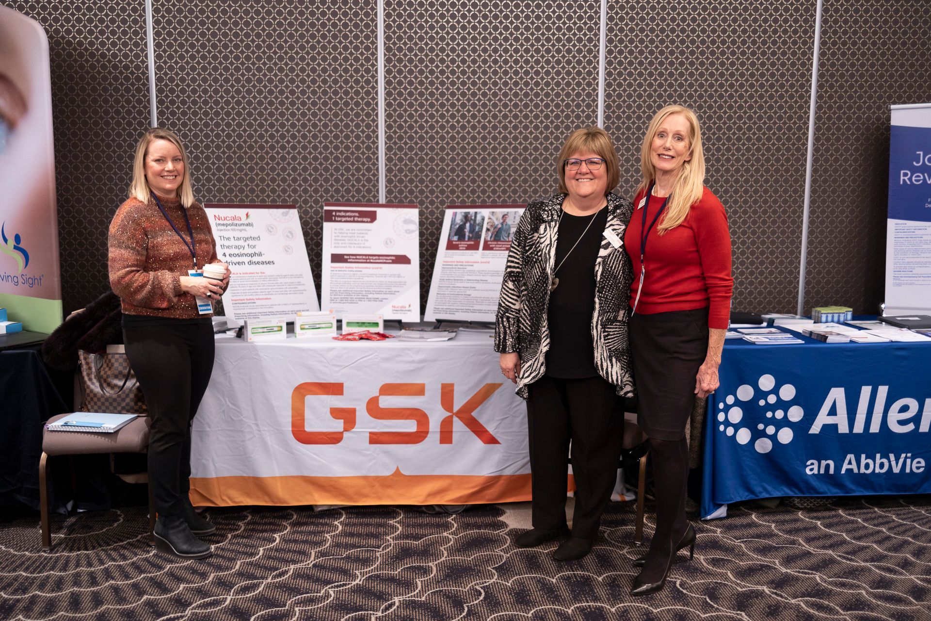 Three women are standing in front of a table that says gsk on it.