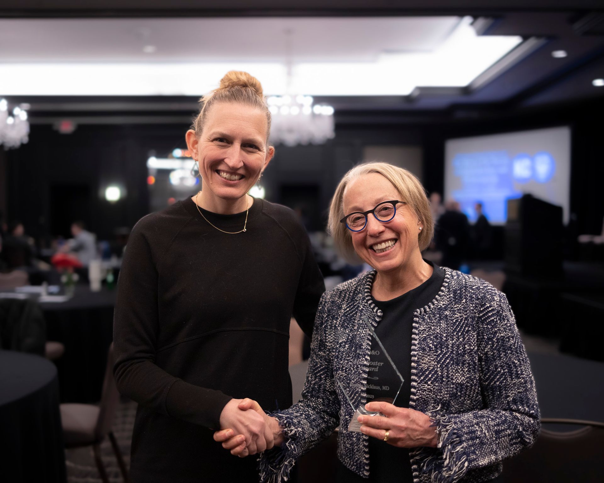Two women are shaking hands in a room and one of them is holding a trophy.