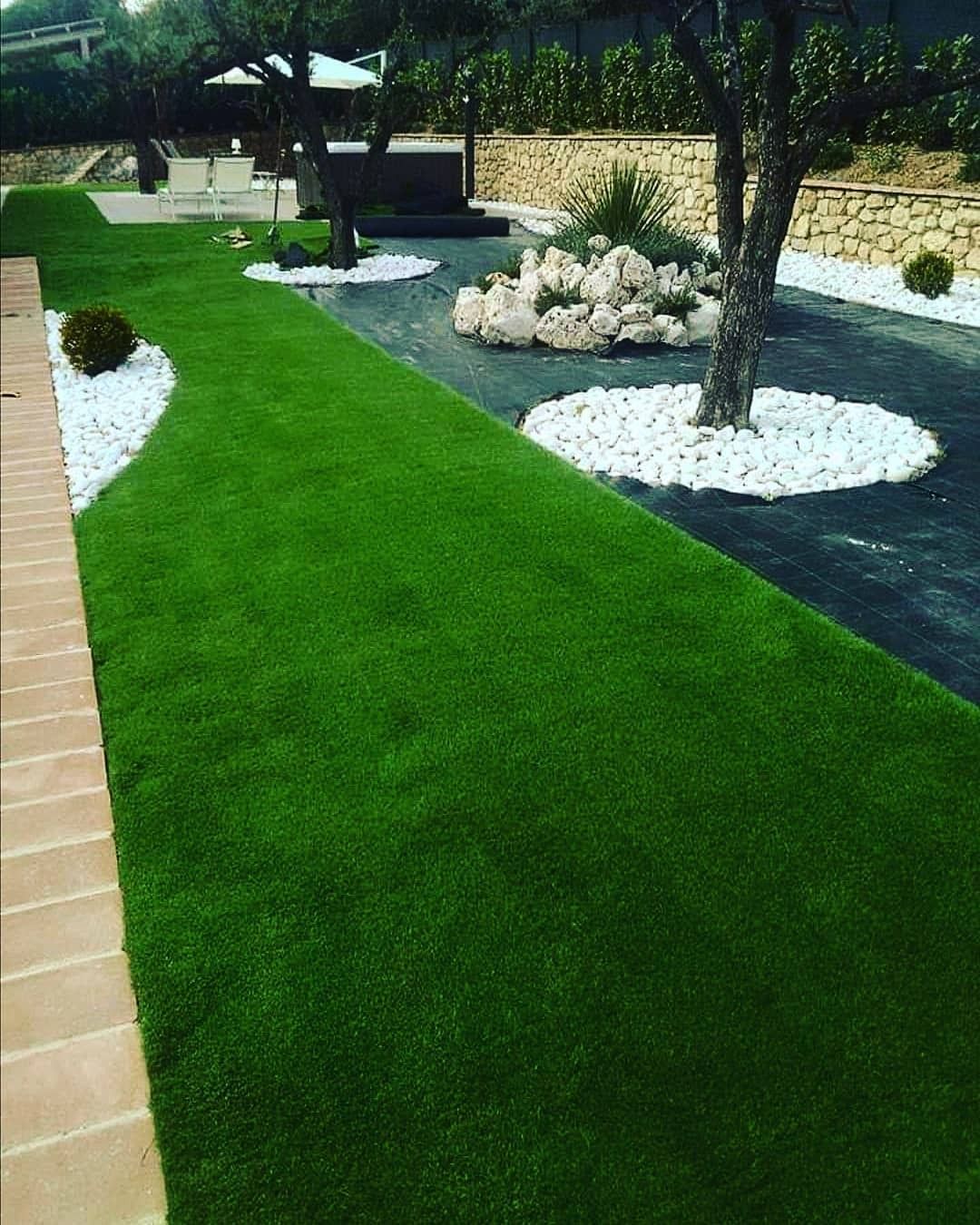 Percorso in erba sintetica verde delimitato da pietre bianche e terracotta, con alberi e patio esterno.