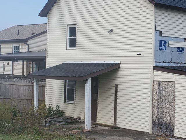 Two-story beige house with a small porch and an attached shed. The roof is dark, and there's a wooden fence.