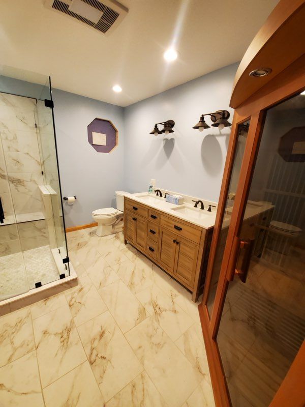 A modern bathroom with a glass shower, double vanity, and a sauna. Light blue walls and marble-patterned floor.