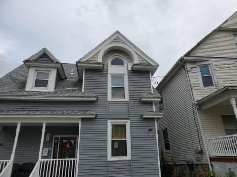 Two-story blue house with porch, dormers, and white trim. Adjacent is a similarly sized house with peeling paint. Cloudy sky.