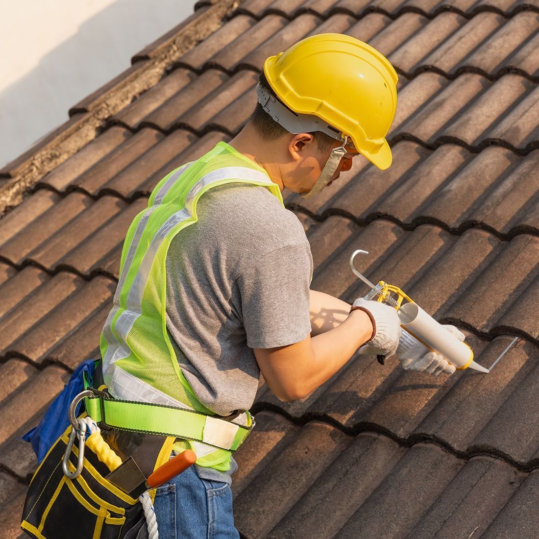 Roofer on a tiled roof, wearing a hard hat and safety vest, applying sealant with a caulk gun.