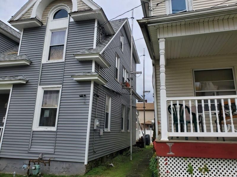 Two multi-story houses; gray siding on the left, white porch with red trim on the right. Narrow alley between.
