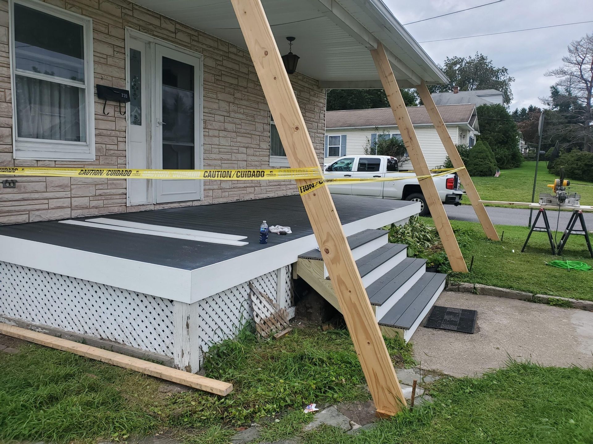 Porch with leaning supports, caution tape, and steps. House with visible damage. Green lawn and a cloudy sky.
