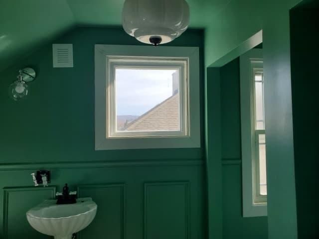 Green-painted bathroom with white trim. Square window overlooking a roof. White pendant light. Sink and mirror.