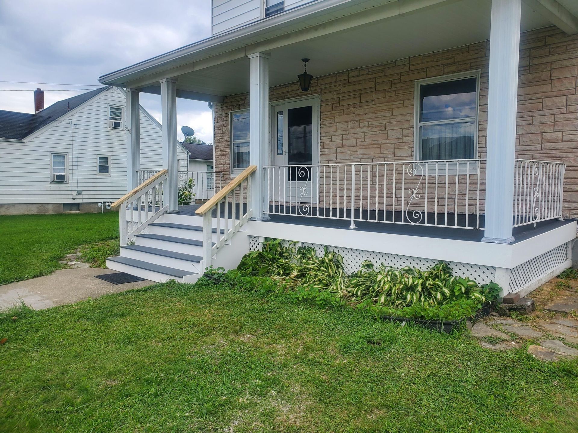 A two-story house with a porch. White railing, stairs, and a door are visible. Green grass and a cloudy sky.