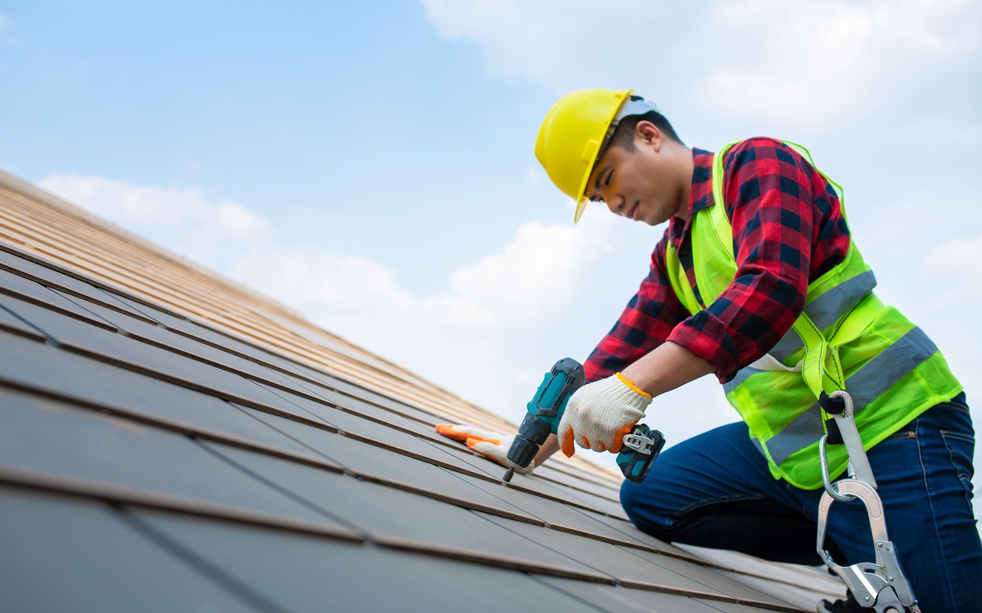 Roofer in yellow hard hat, safety vest, and gloves, using a drill on a tile roof against a blue sky.