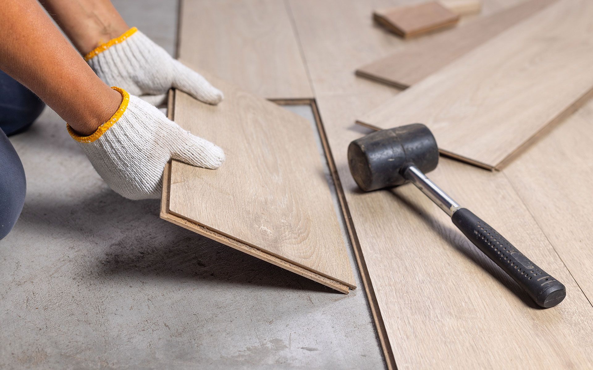 Person wearing gloves fitting a laminate wood floor plank with a mallet nearby.