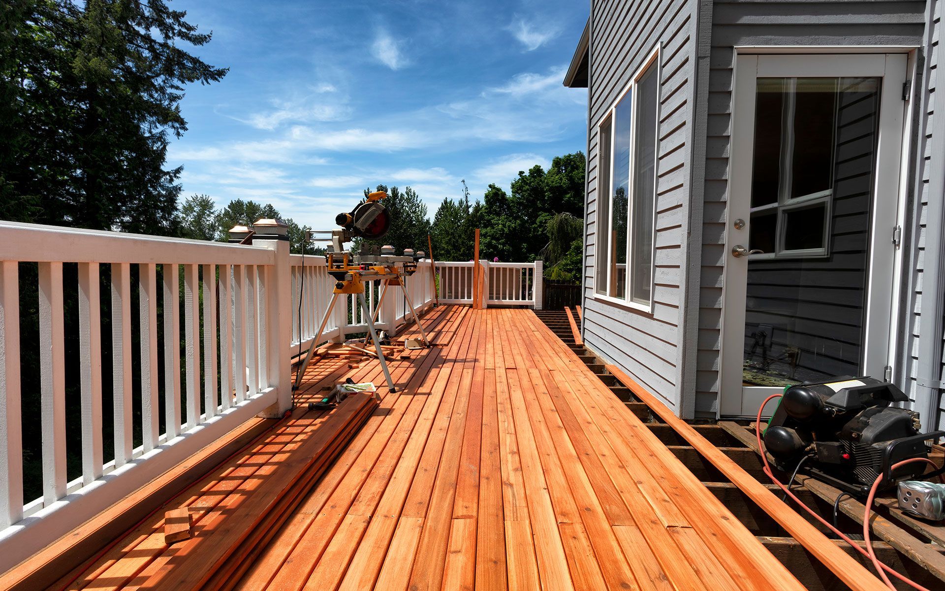 Newly constructed wood deck with white railing, against a gray house, under a blue sky.