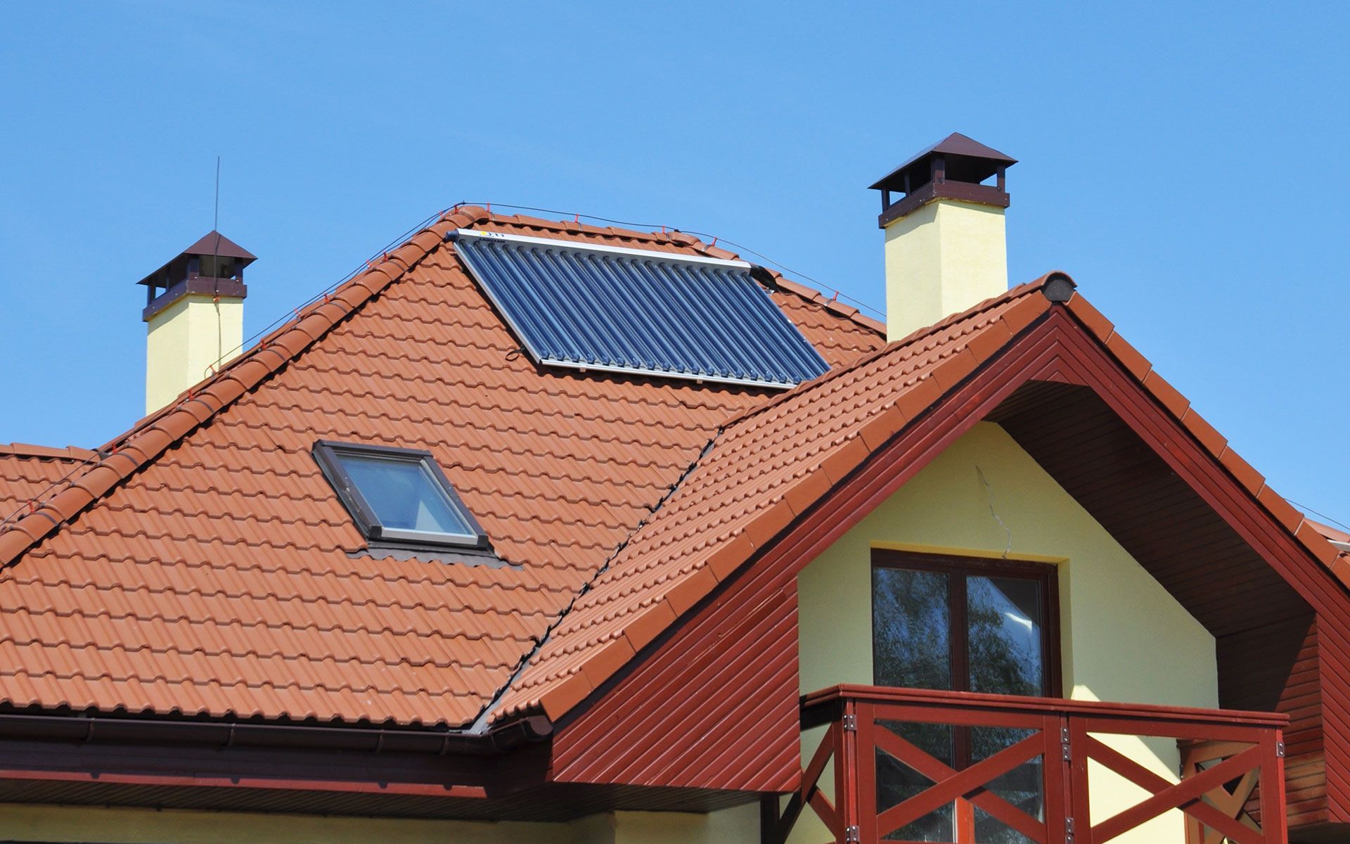 A house with a red tiled roof, solar panels, chimneys, and a small balcony against a blue sky.