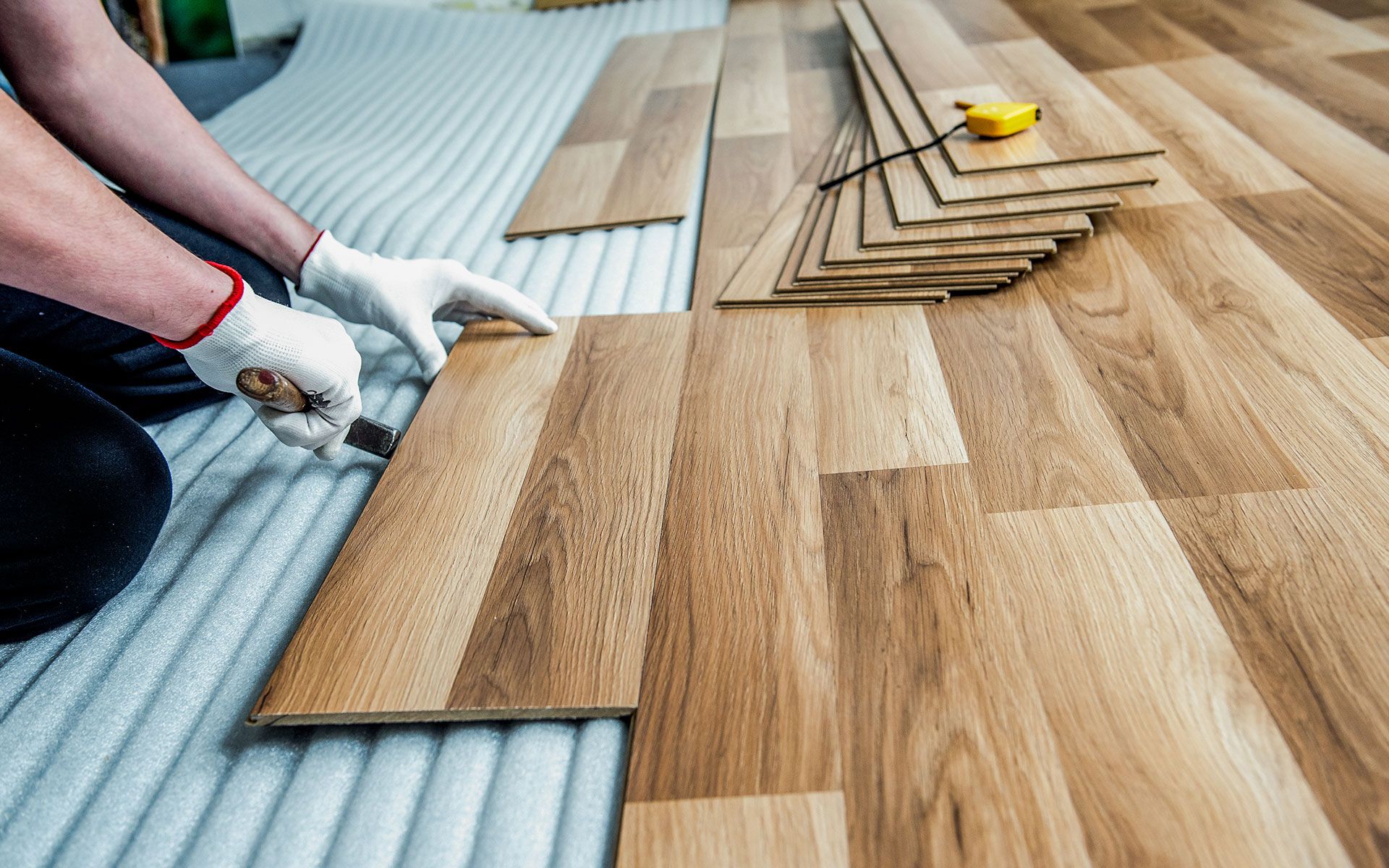 Person installing wood flooring, wearing gloves, using a knife.