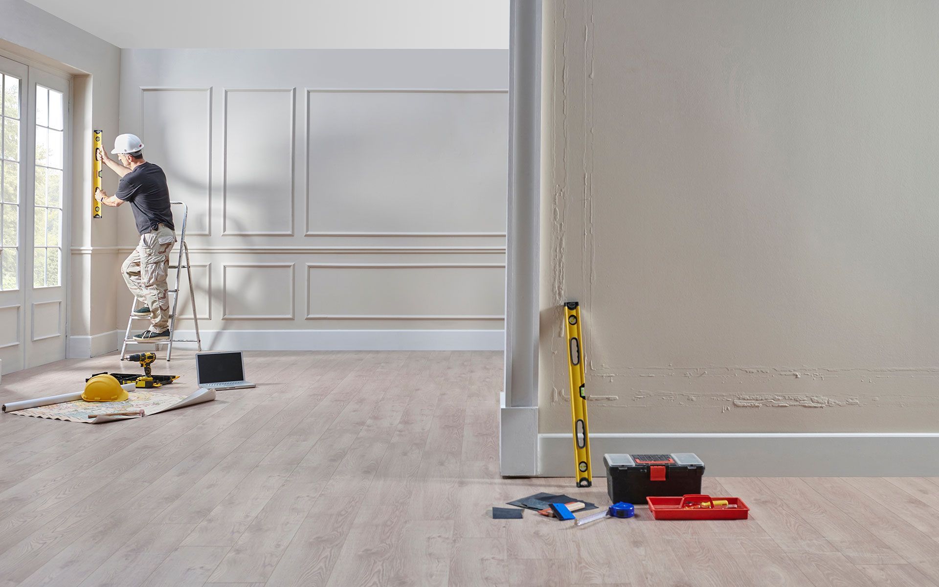 Man using a level in a room with tools on the floor; molding detail, light wood floor, bright room.