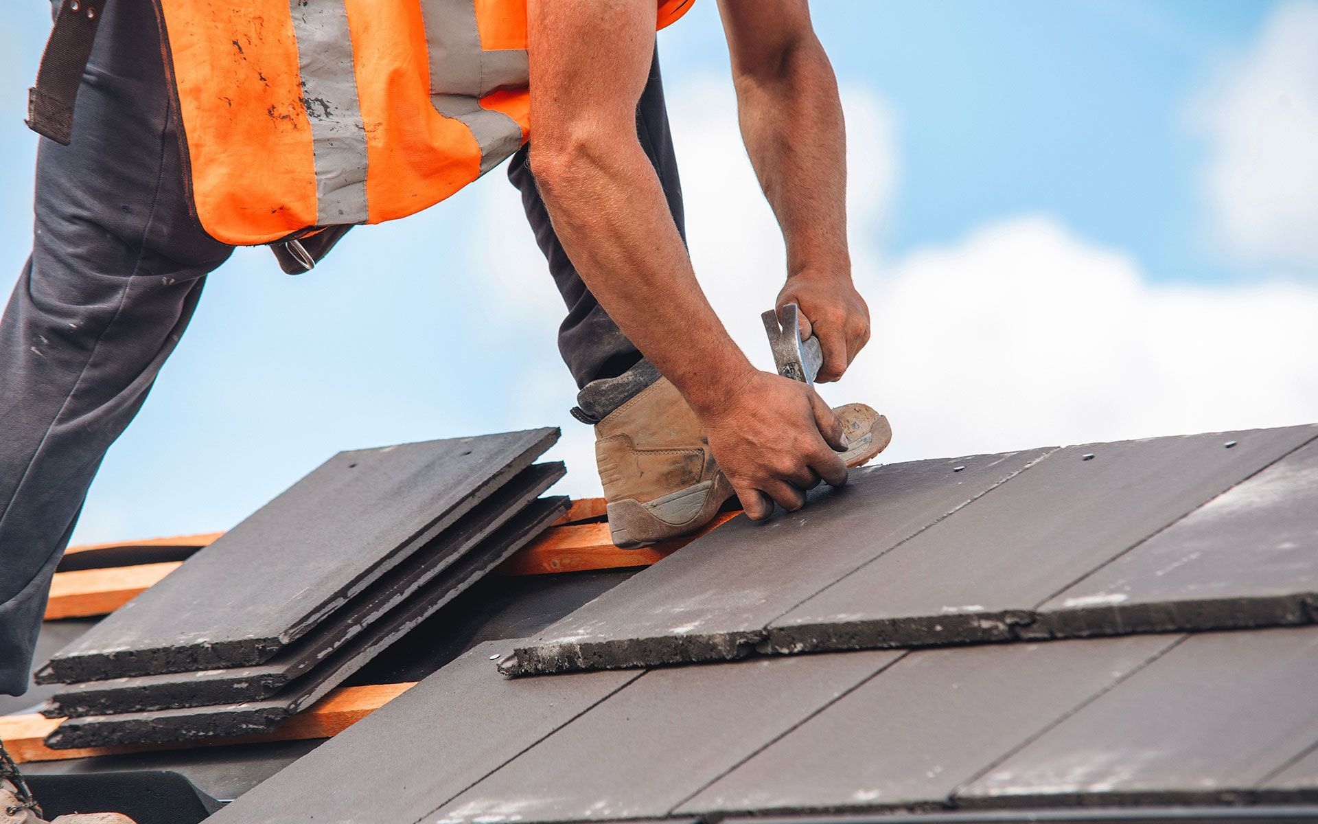 Roofer installing dark gray tiles on a roof, wearing an orange safety vest, under a blue sky.