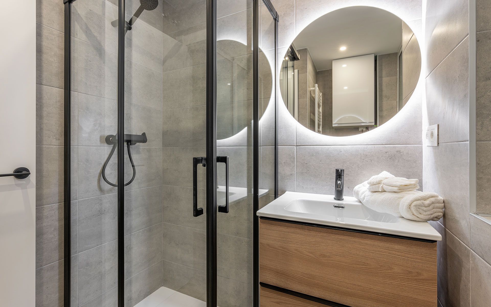 Modern bathroom with a glass shower, wooden vanity, and a round illuminated mirror.