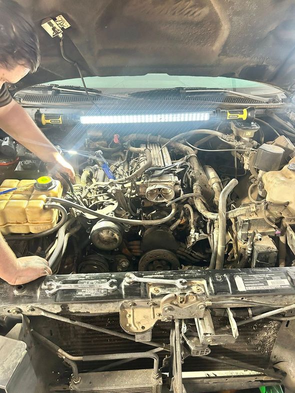Mechanic working on a car engine under bright LED light. Engine bay is dirty.