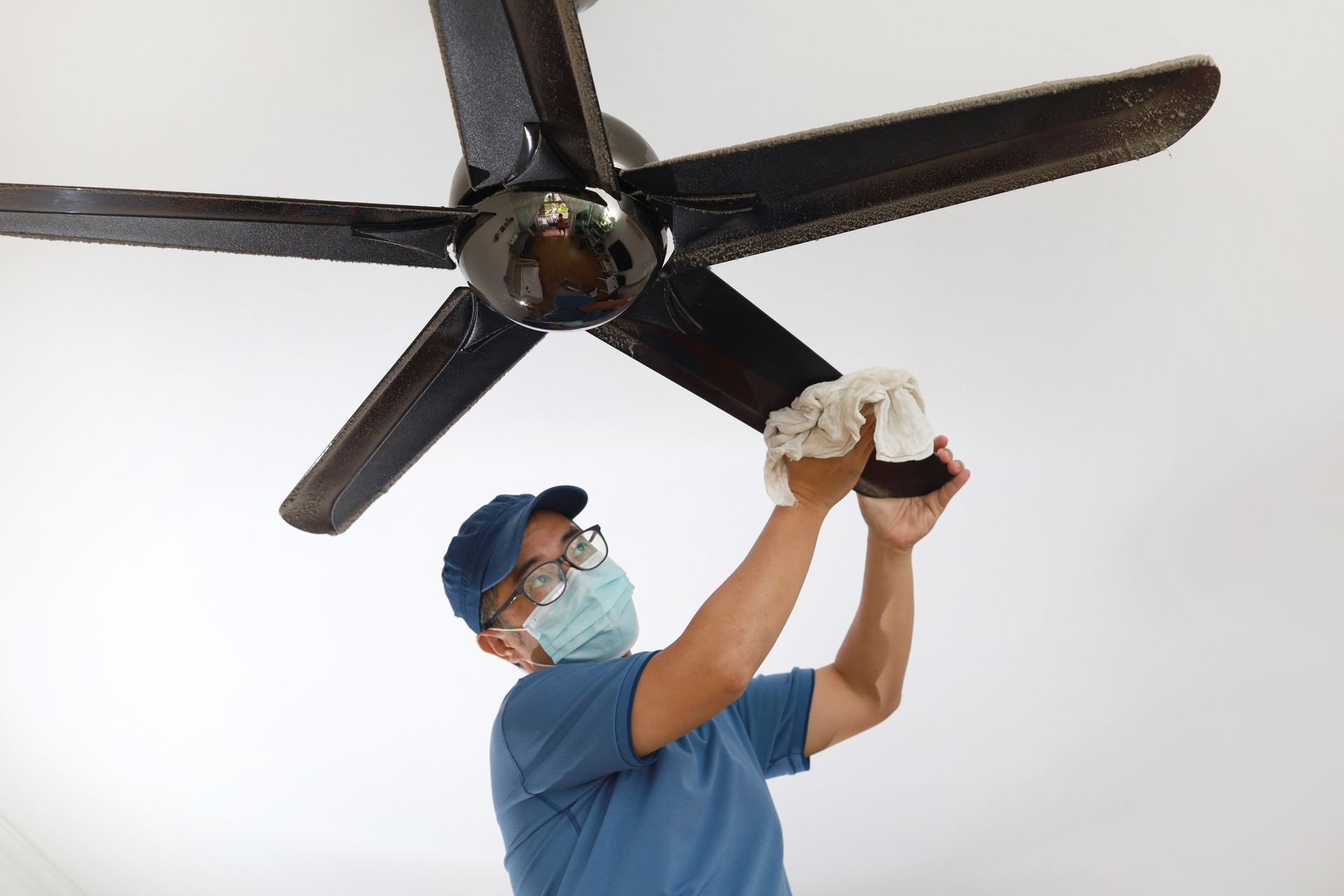 A man wearing a mask is cleaning a ceiling fan.