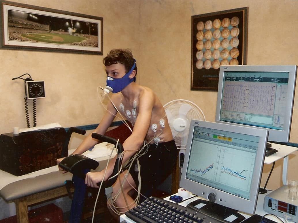 Person wearing a mask and sensors, cycling indoors with monitors displaying data.