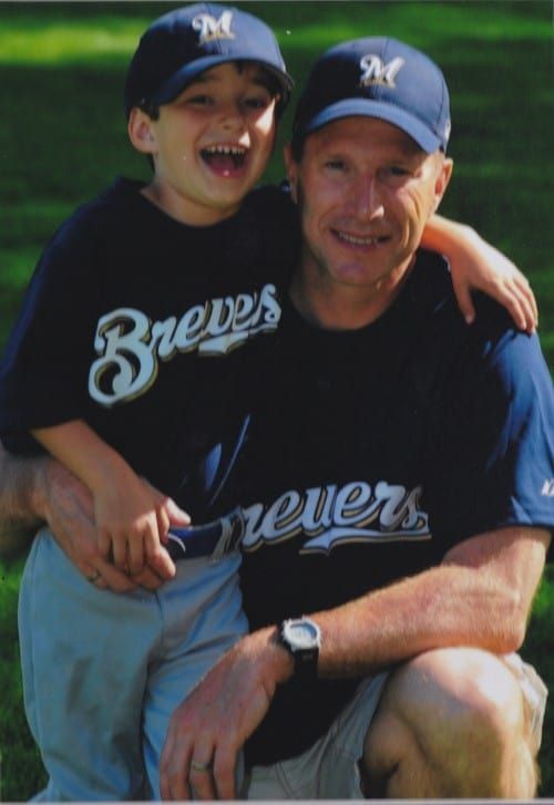 Man and child in matching Brewers baseball shirts and hats, posing outdoors. Child has an arm around the man.