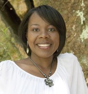 Woman with black hair, smiling, wearing a white top and necklace, outdoors.