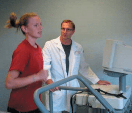 Woman running on treadmill, doctor observing, medical equipment in background.