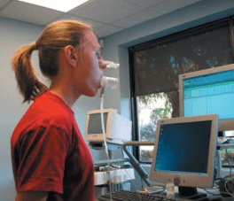 Woman in red shirt testing breath with tubes in mouth, monitors in background.