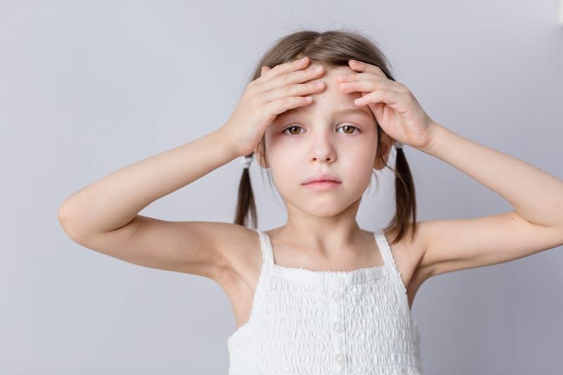 Dysautonomia symptoms. Girl in a white top with her hands on her forehead, looking concerned.