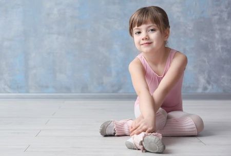 Girl in pink leotard and ballet shoes sits on floor, smiling. Blue and gray background.