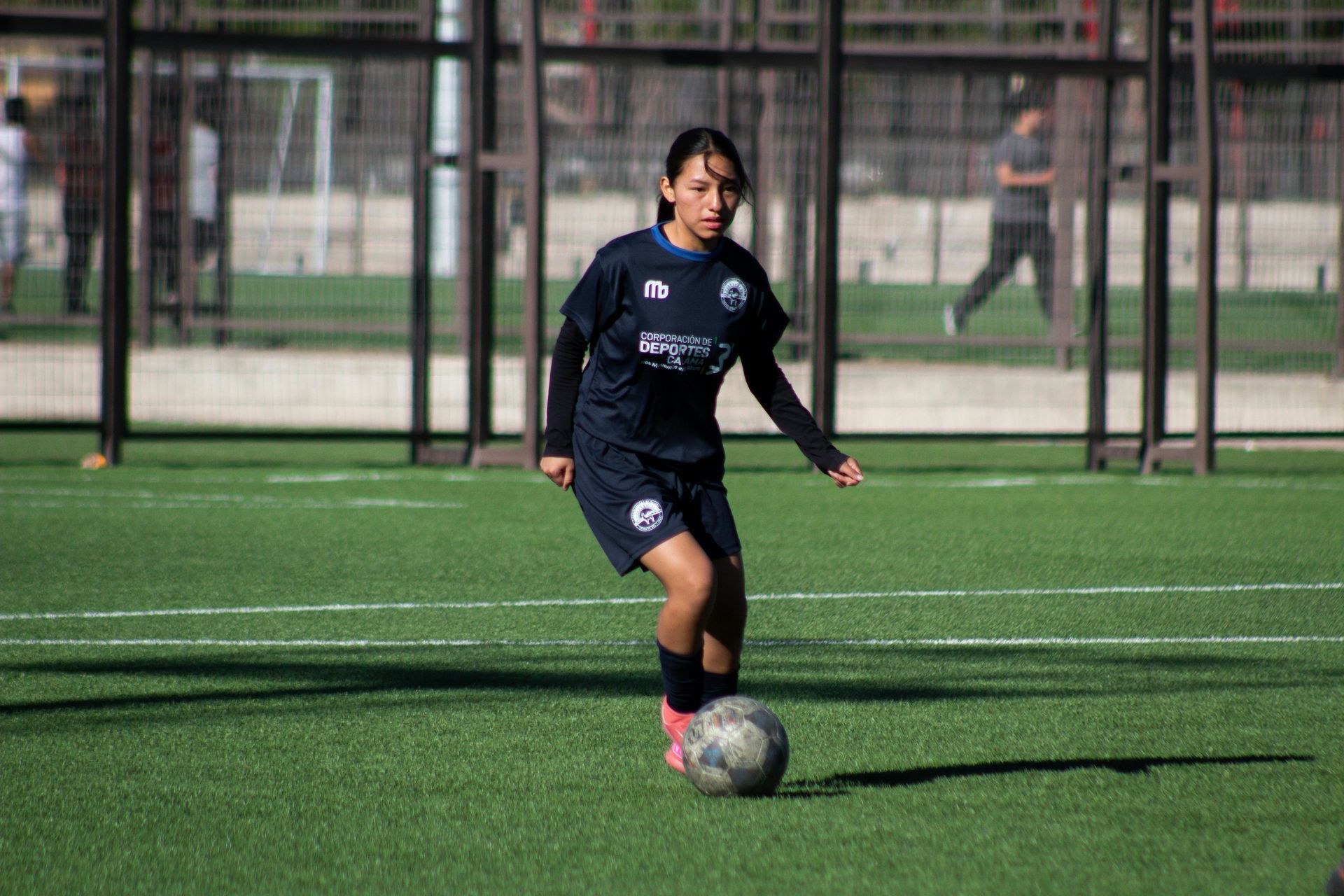 Soccer player in blue uniform dribbling a ball on a green turf field, outdoor setting.