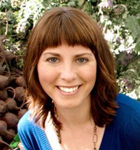 Woman with shoulder-length brown hair, bangs, smiling, wearing blue sweater, against leafy background.