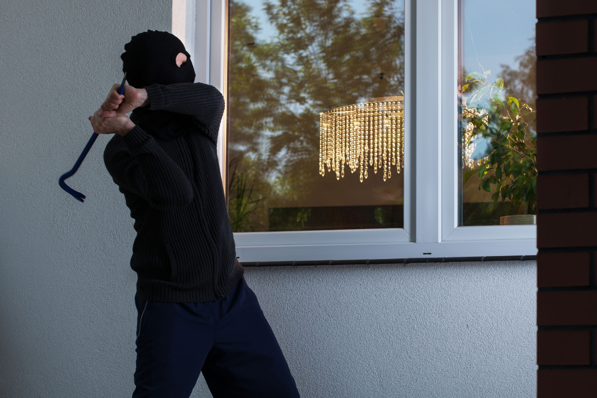 A man in a mask attempting to break into a house through a window, showing the importance of security tint.