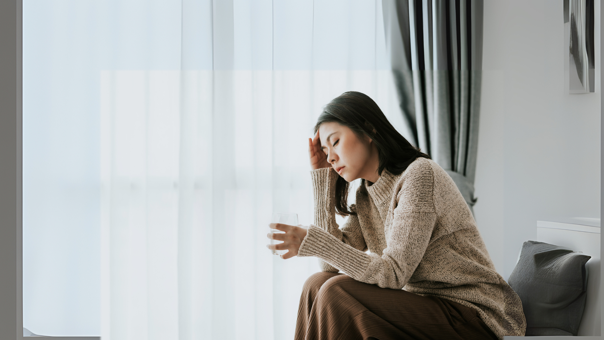 Woman with a headache, holding a glass, sitting by a window, looking distressed.