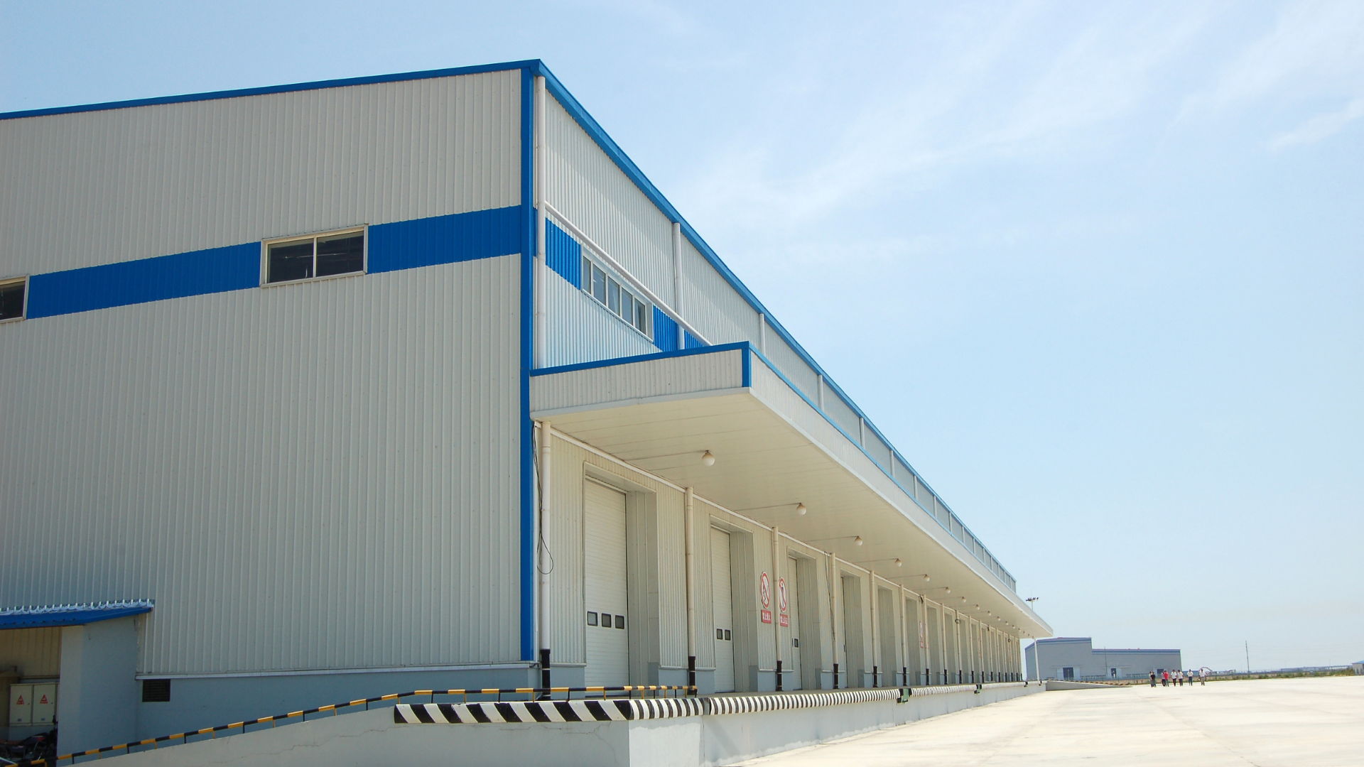 Large, white industrial building with blue trim and loading docks under a bright sky.