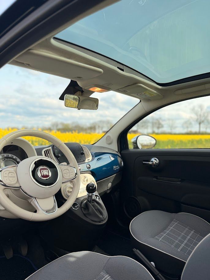 Witte Fiat 500 met beige dashboard, blauwe accenten en schuifdak, geparkeerd bij gele velden.
