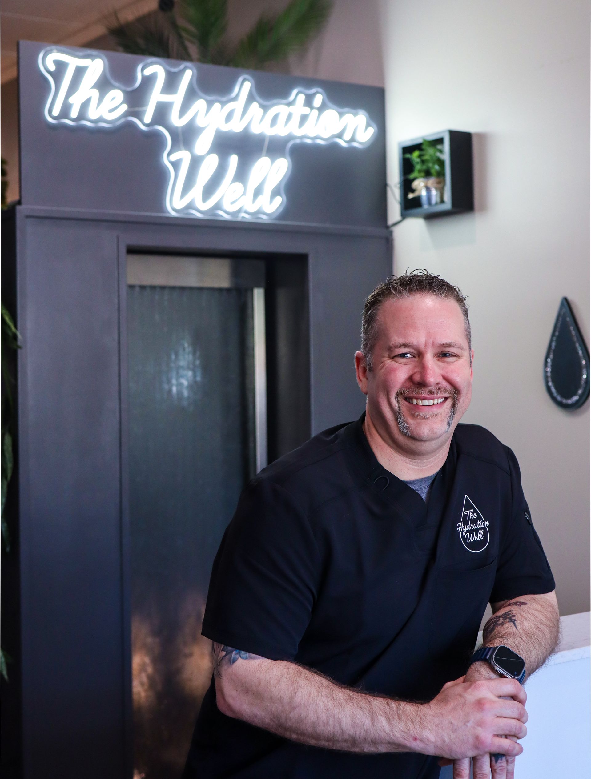Smiling person standing at The Hydration Well with a neon sign on the wall behind them