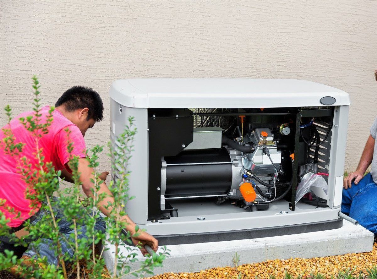 Two people installing a gray generator near a beige wall, outdoors. One person is in pink, the other in blue.