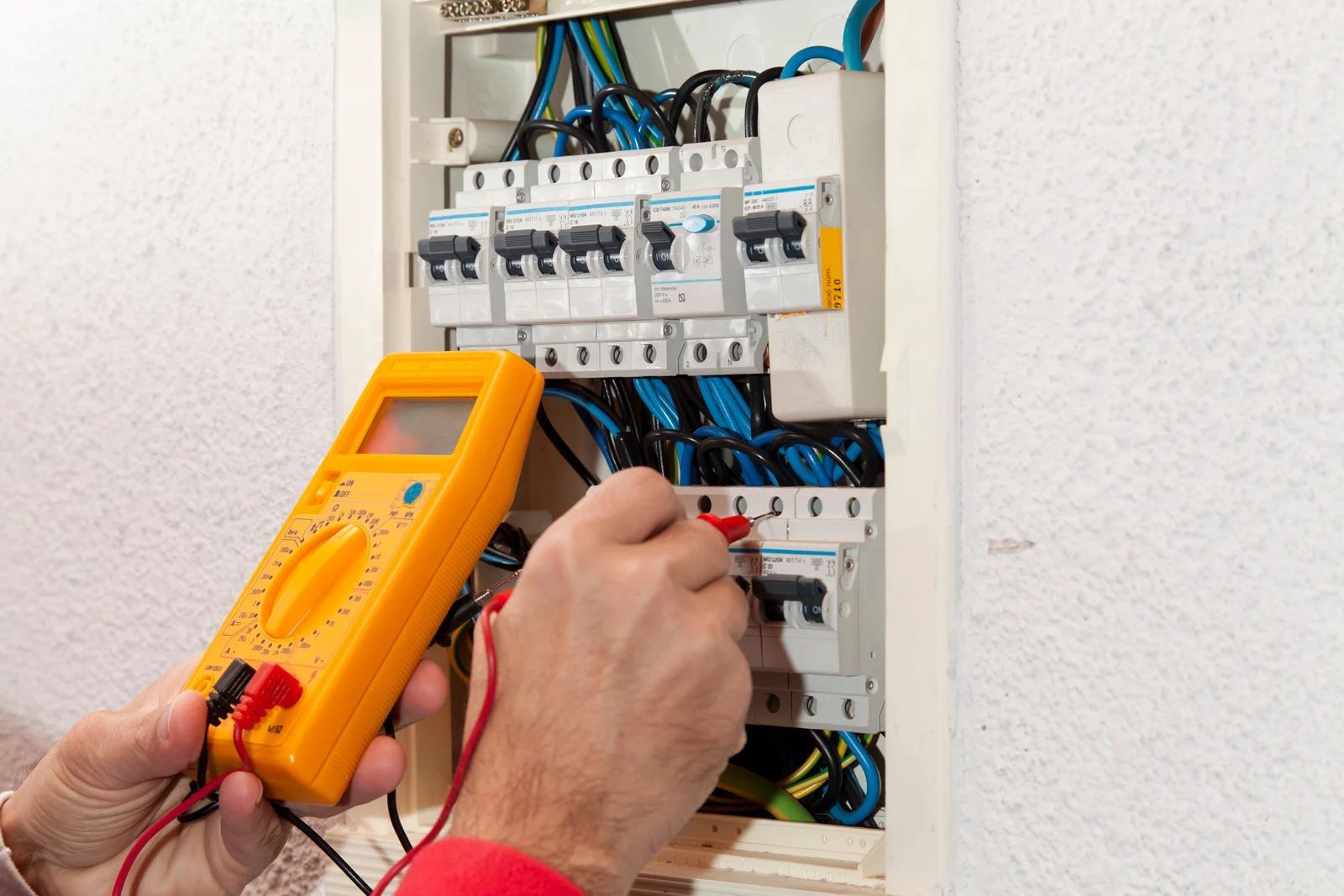 A person using a yellow multimeter to test a circuit breaker panel, indoors.