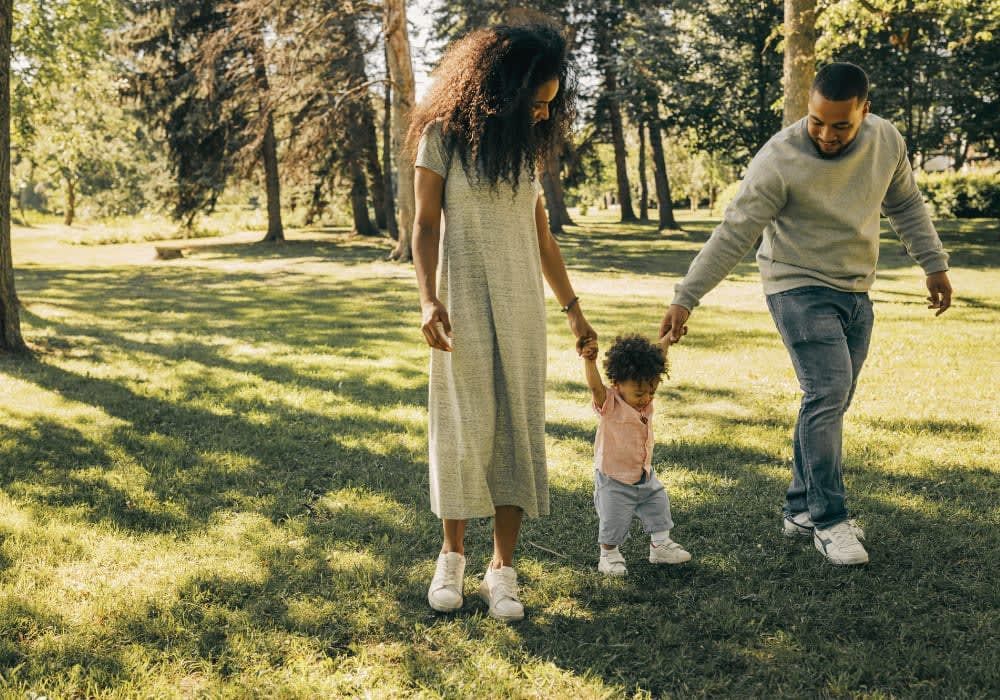Family walking in a sunny park: mother holding child's hand, father beside them. Green grass, trees.