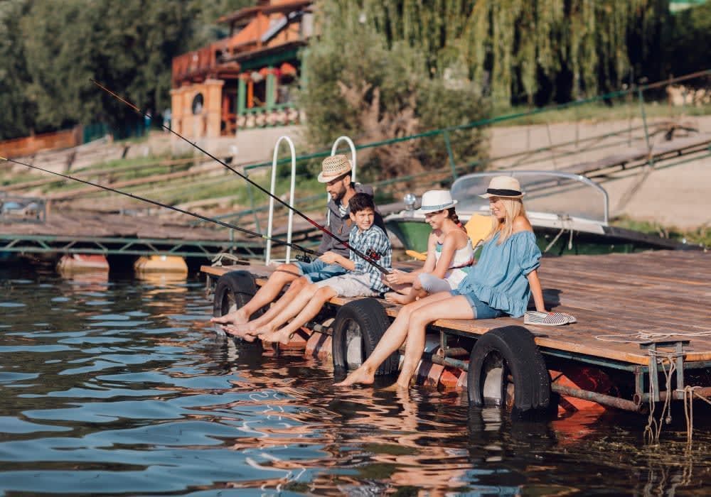 Family fishing on a dock, feet dangling in water. Wooden dock, lake, sunny day, house in the background.
