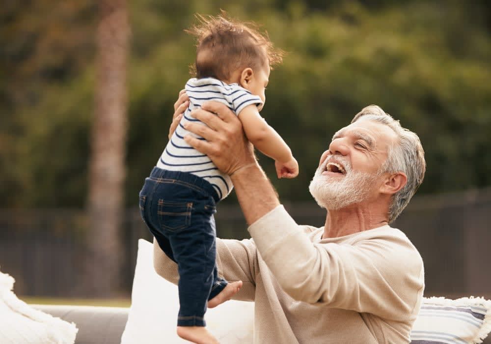 A smiling grandfather holds a baby up in the air outside.