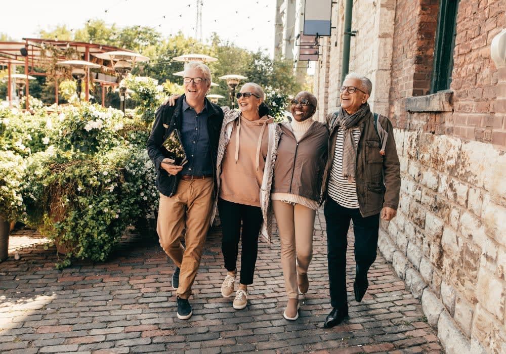 Four smiling seniors walking arm-in-arm on a brick path near a brick building and green shrubs.