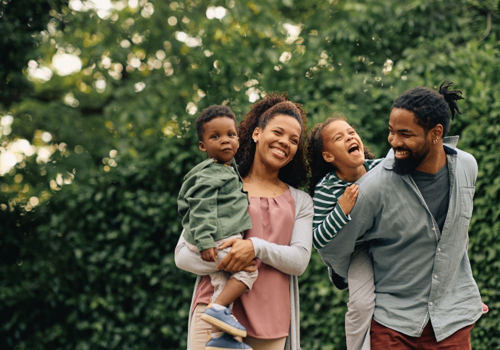 Family of four smiles outdoors; mom holds toddler, dad carries child piggyback. Green backdrop.