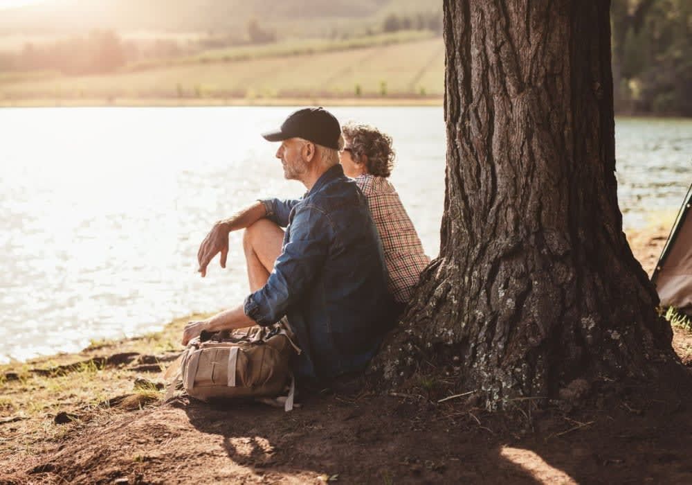 Couple sits by a lake under a tree. Man wears cap, looks away, woman rests against him.
