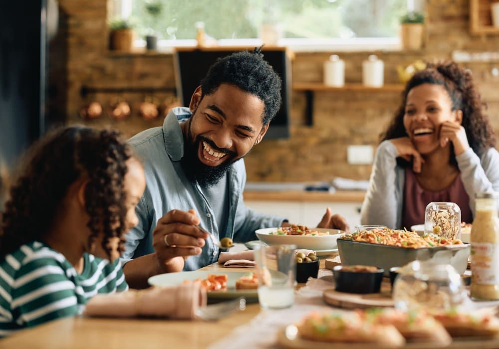 Family laughing and eating together at a wooden table in a bright kitchen.