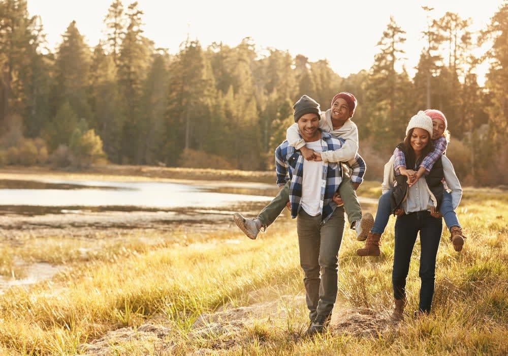 Family hiking, piggybacking in front of a lake and trees on a sunny day.