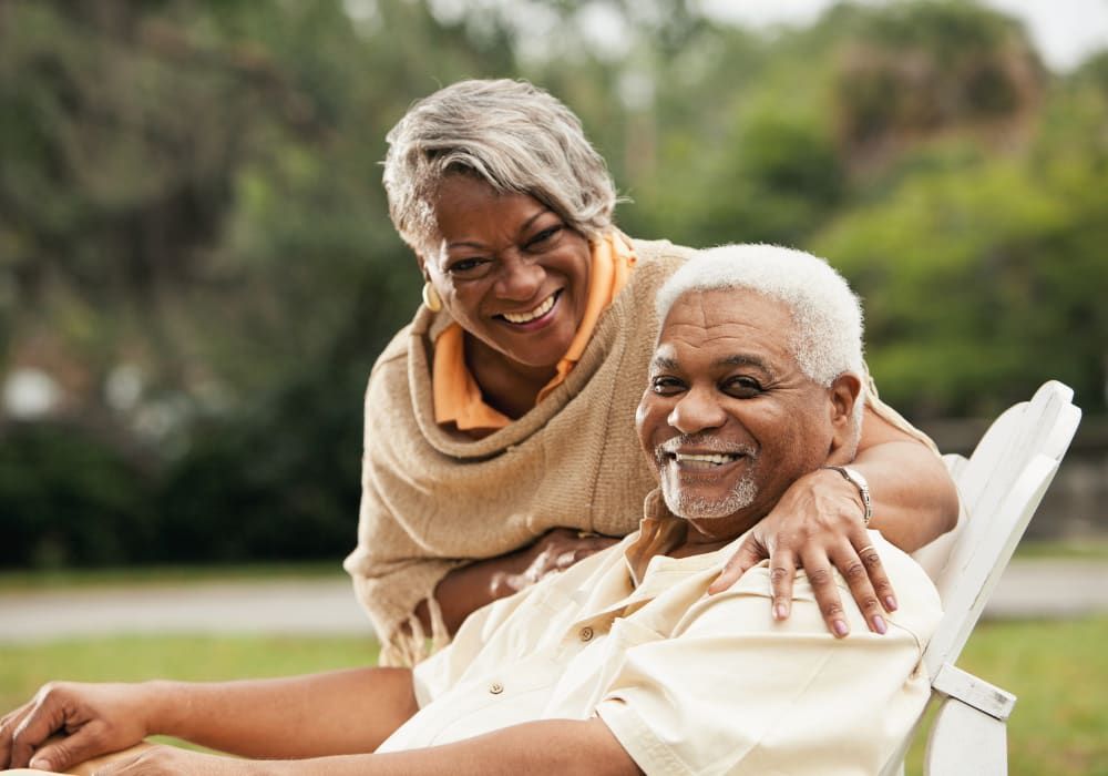 Smiling elderly couple, woman's arm around man seated in a chair outdoors, green background.