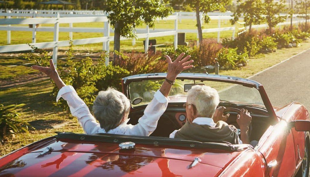 Elderly couple in red convertible with arms raised, driving down road with white fence.
