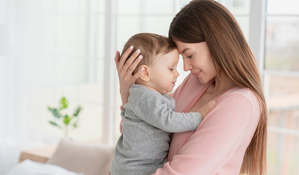 A young woman in a pink shirt embraces a baby, forehead to forehead, near a window in a well-lit room.